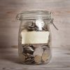 40278822 - coins in glass money jar with blank label, financial concept. vintage wooden background with dramatic light.